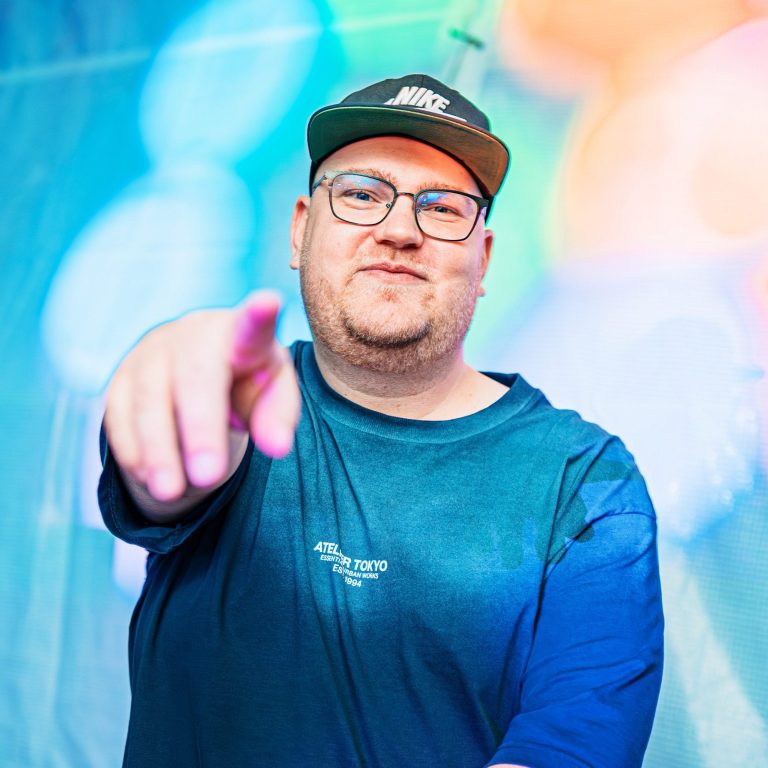 DJ Driessen zeigt mit dem Finger auf die Kamera, farbenfroher Hintergrund.