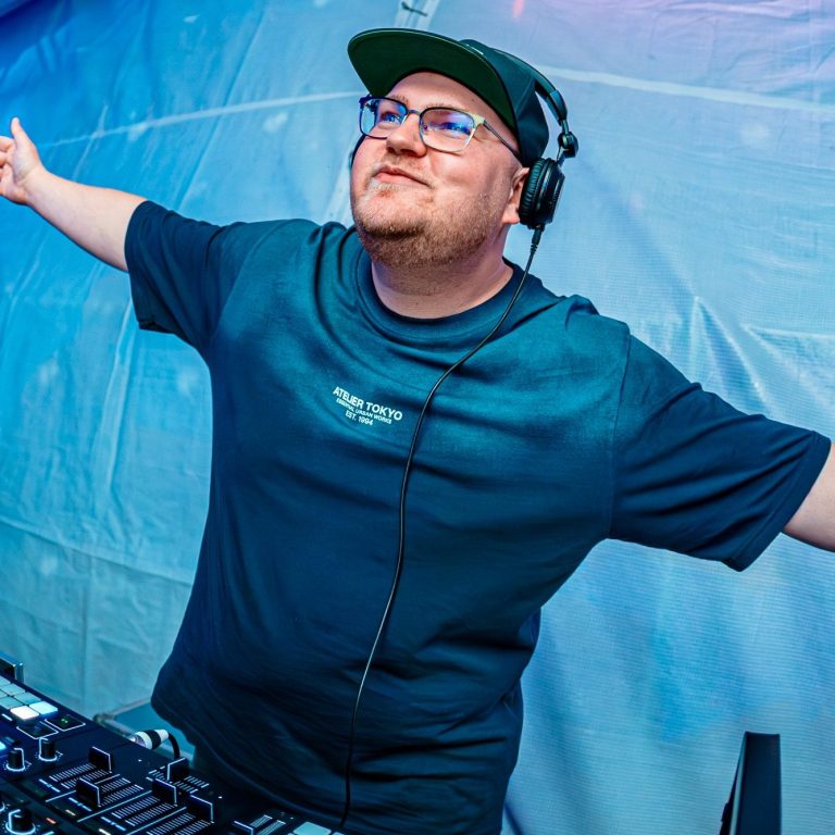 DJ Driessen steht hinter einem Mischpult und hebt die Arme in die Luft, während er auflegt.