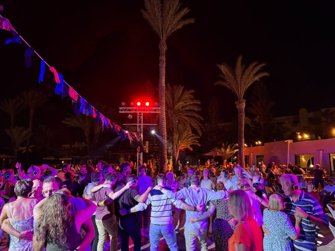 Menschen tanzen und feiern unter Palmen bei Nacht, beleuchtet von bunten Lichtern.