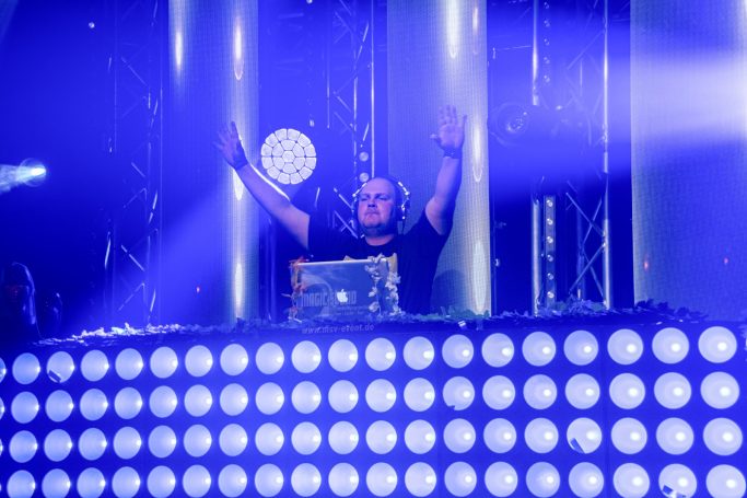 DJ steht an einem Mischpult, beleuchtet von bunten Lichtern, Hände in die Luft.