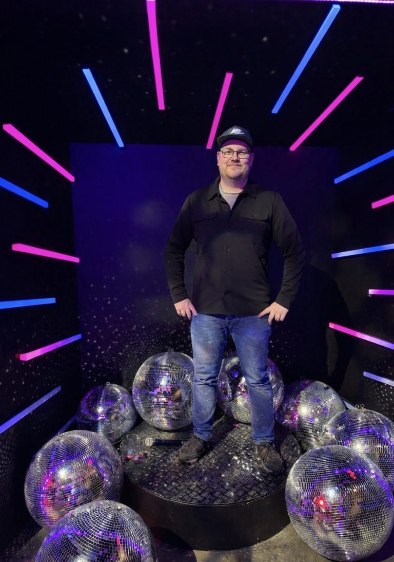 DJ Driessen steht auf einer Plattform umgeben von spiegelnden Kugeln und bunten Lichtern.