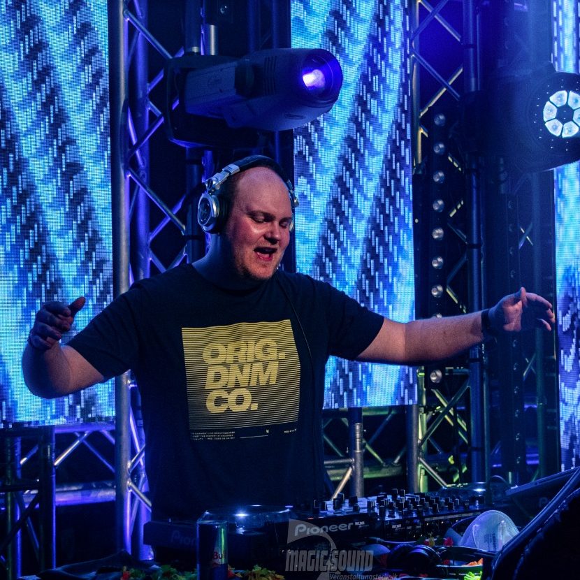 DJ Driessen mit Kopfhörern, der auf einer Festival Bühne zu Musik auflegt, umgeben von bunten Lichtern.