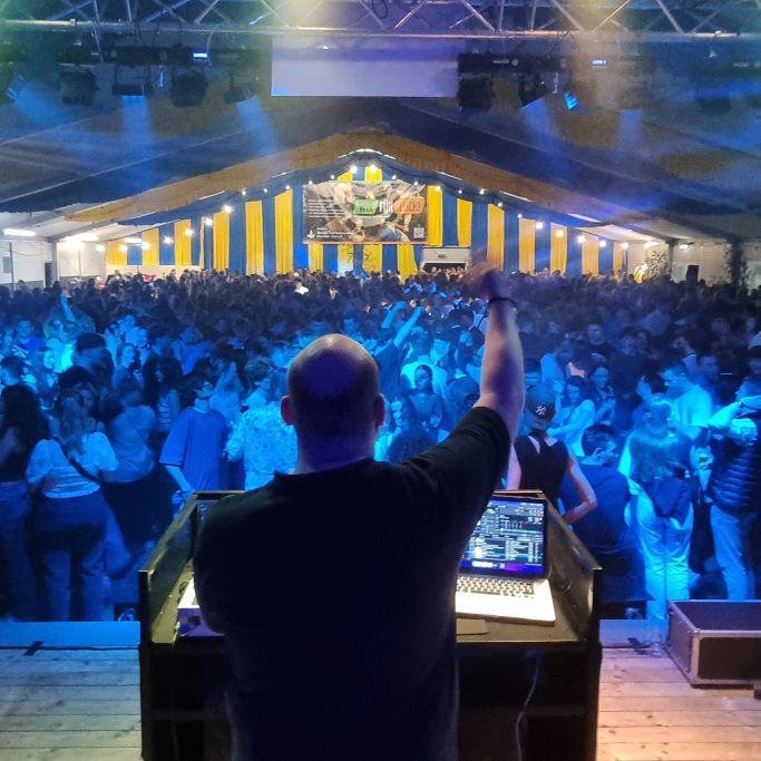 DJ präsentiert vor einer tanzenden Menge in einer festlich beleuchteten Umgebung.
