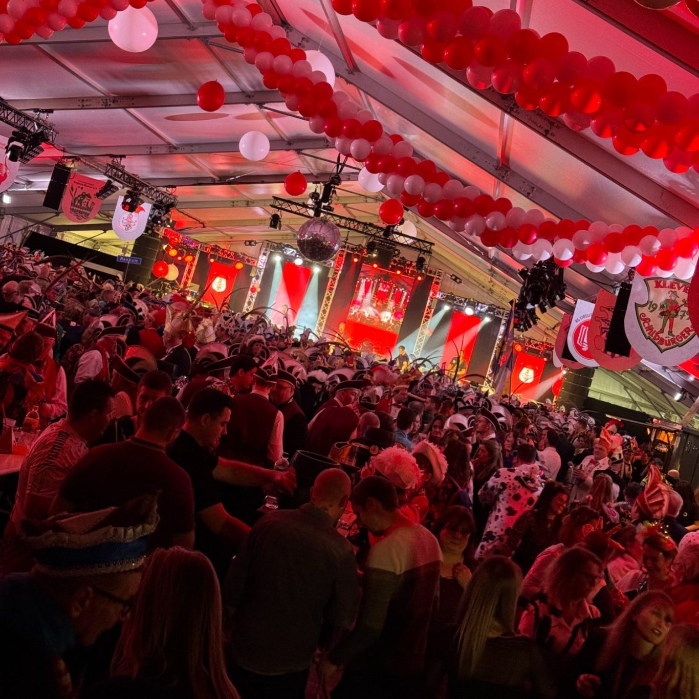 Volles Zelt mit Rot-Weißen Dekorationen, Menschen feiern und tanzen. DJ Driessen auf einer großen Bühne.