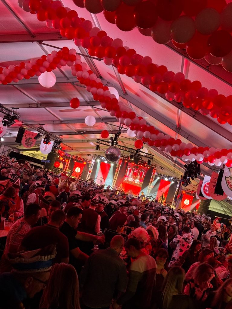 Bunte Party mit vielen Menschen, roten Ballons und festlicher Beleuchtung in einem Zelt.