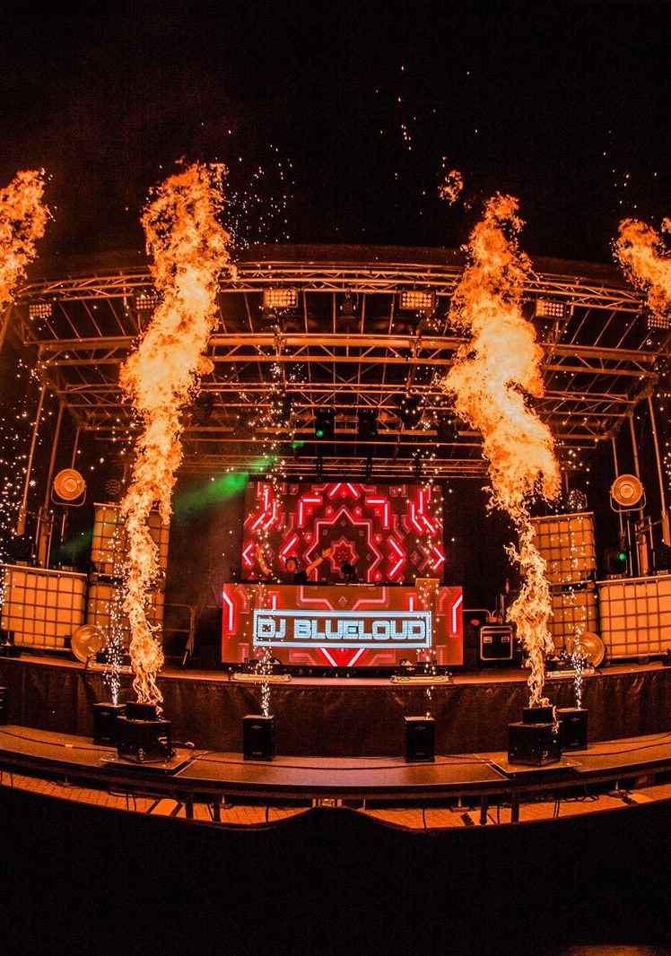 Bühne mit DJ Driessen, bunten Lichtern und Feuerfontänen bei Nacht.