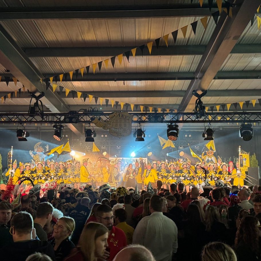 Bunte Karnevalsfeier mit Menschenmengen und gelben Fahnen in einer festlichen Halle.