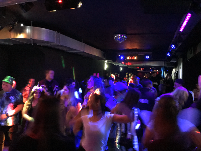 Menschen tanzen in einem dunklen Club, farbige Lichter blitzen im Hintergrund.